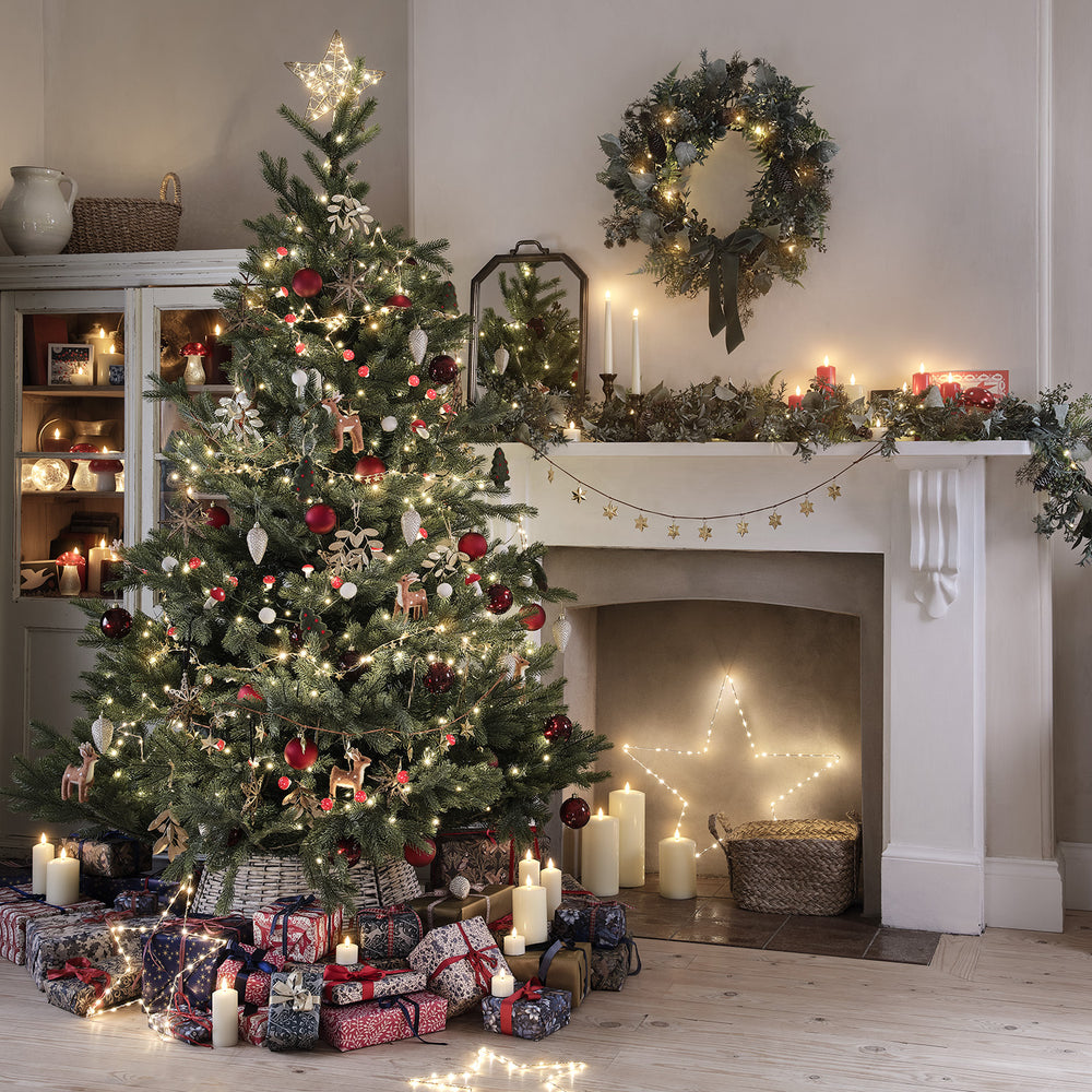 Weihnachtsbaum mit roten Kugeln und Lichterkette auf grauem Rattan Baumrock vor Kamin