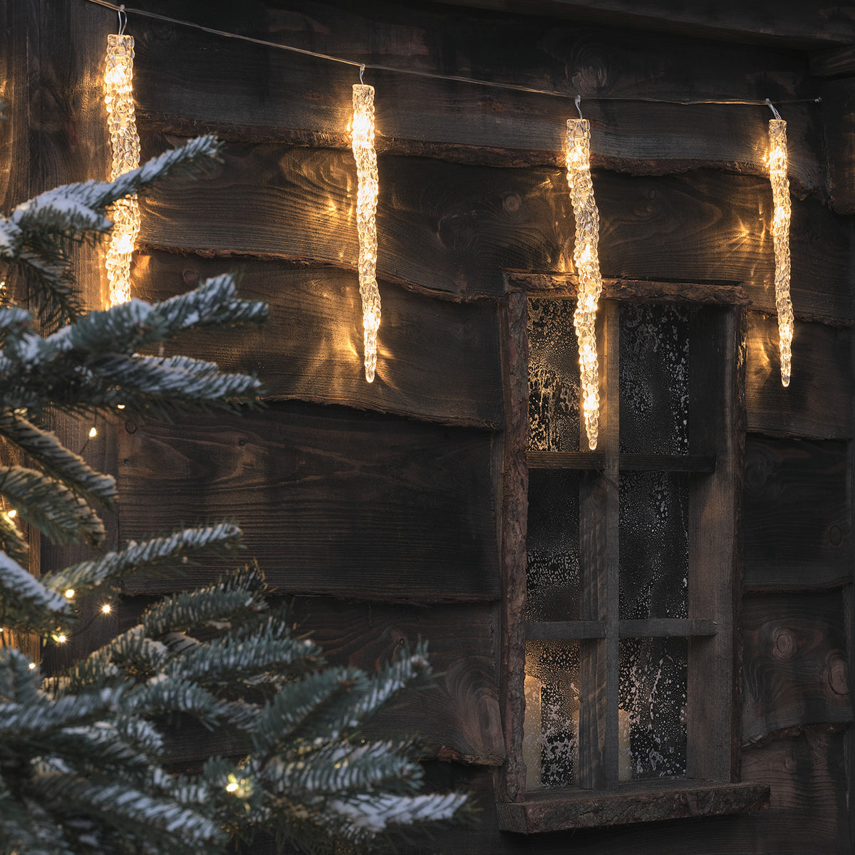 LED Eiszapfen-Lichterkette an Holzhaus mit verschneitem Tannenbaum im Vordergrund