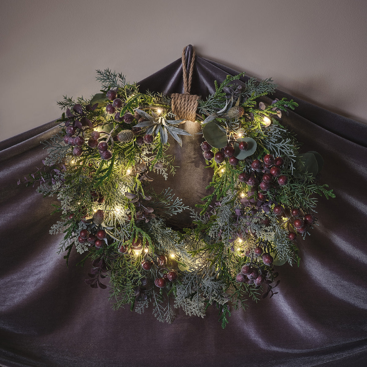 Beleuchteter Weihnachtskranz mit lila Beeren, Tannenzapfen und Juteschlaufe vor dunklem Stoff
