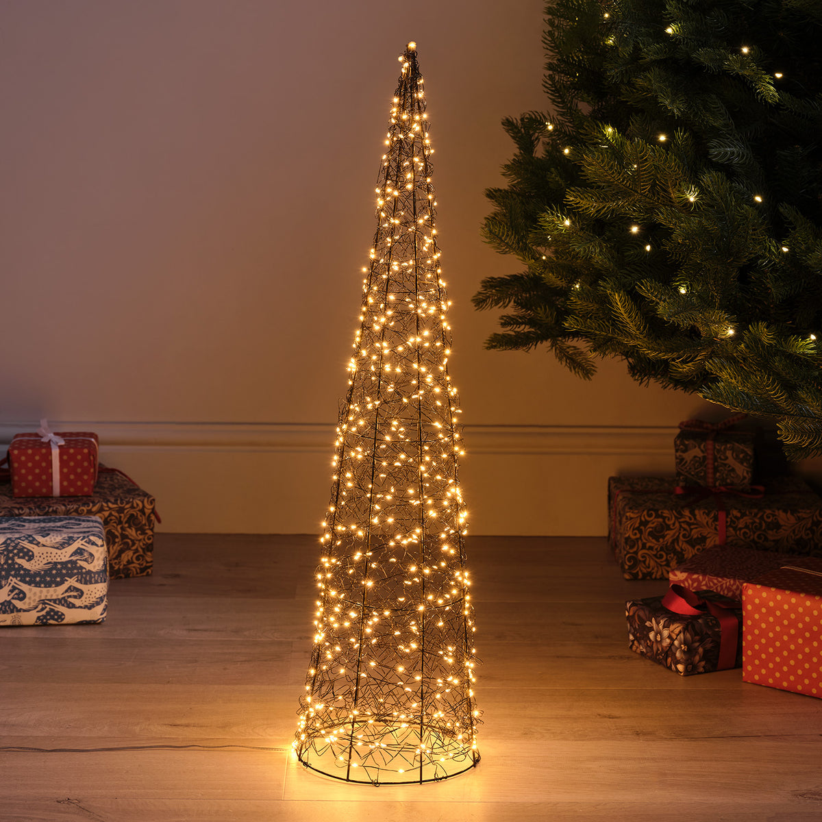 Großer LED-Kegel aus silberfarbenem Metalldraht mit warmweißen Micro-LEDs neben Weihnachtsbaum und Geschenken