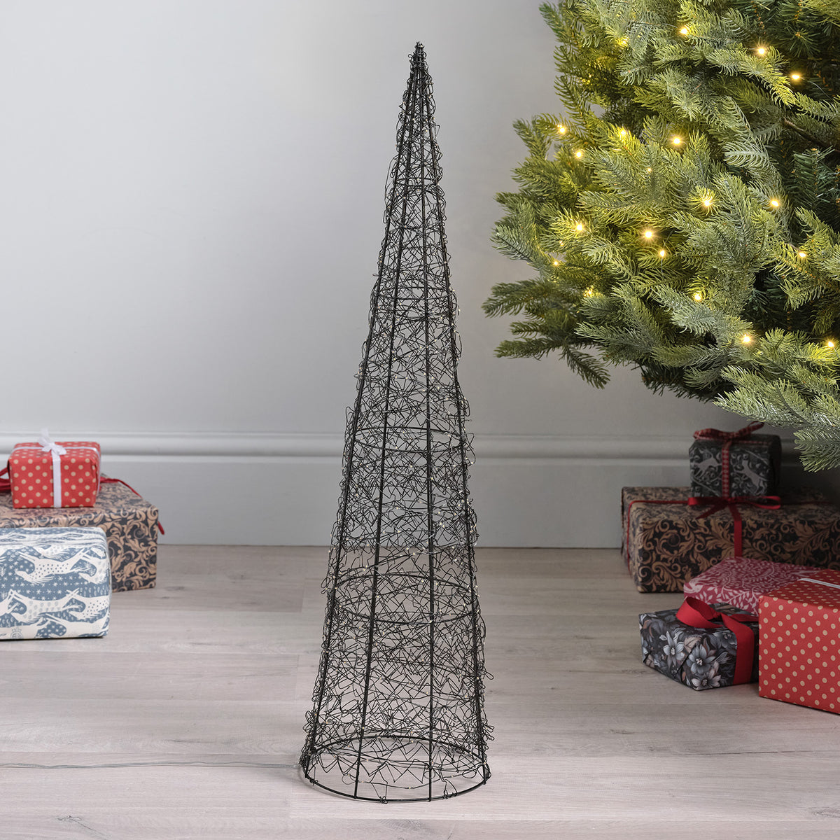 Unbeleuchteter kleiner LED-Kegel aus Metalldraht vor Weihnachtsbaum und Geschenken auf Holzboden