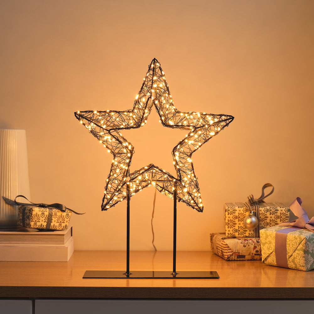 Leuchtender 3D-Stern aus schwarzem Draht mit warmweißen Micro-LEDs auf Metallständer vor warmem Hintergrund