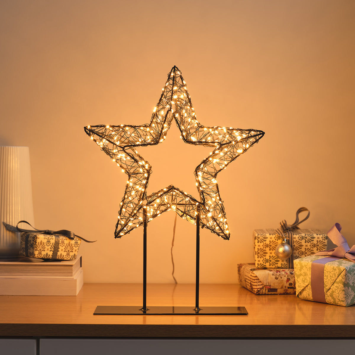 Leuchtender 3D-Stern aus schwarzem Draht mit warmweißen Micro-LEDs auf Metallständer vor warmem Hintergrund