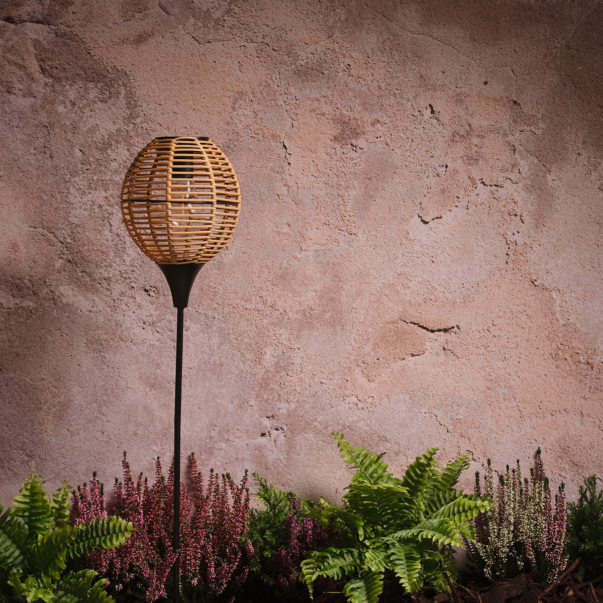 Solar-Gartenstecker mit rundem Rattan-Schirm auf Metallstab vor strukturierter Wand und Pflanzen