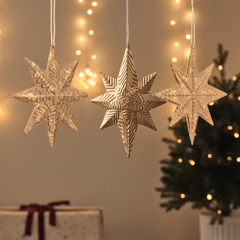 Goldene Papierstern-Weihnachtsanhänger vor beleuchtetem Fenster, geschmückter Tannenbaum mit Geschenk im Hintergrund.
