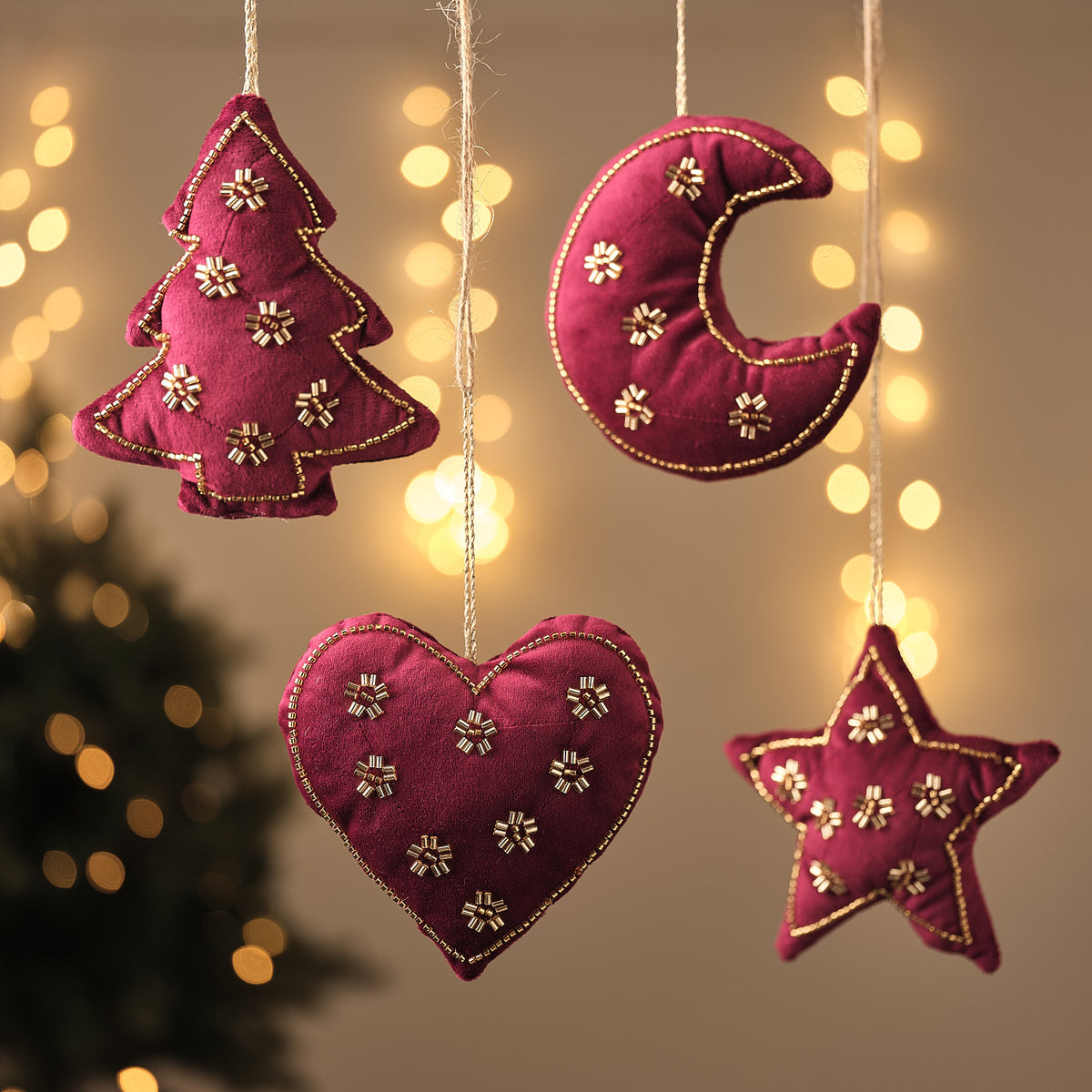 Bordeauxrote Stoff-Weihnachtsanhänger in Tannenbaum-, Mond-, Herz- und Sternform mit goldener Blumenstickerei, hängend vor Lichterglanz.