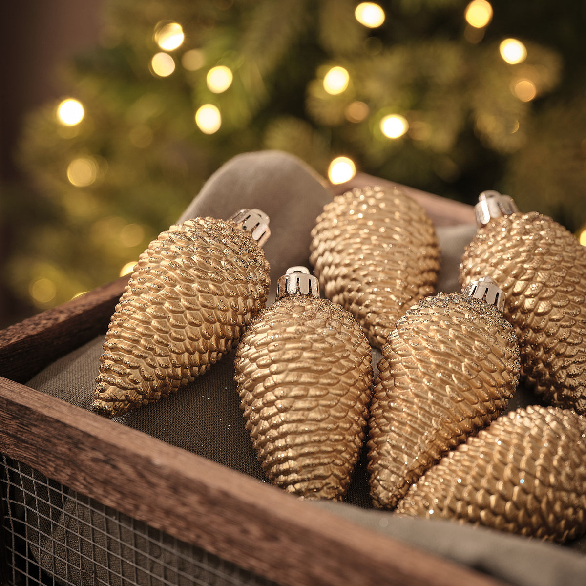 Goldene Tannenzapfen-Weihnachtskugeln in Holzkiste vor beleuchtetem Tannenbaum.