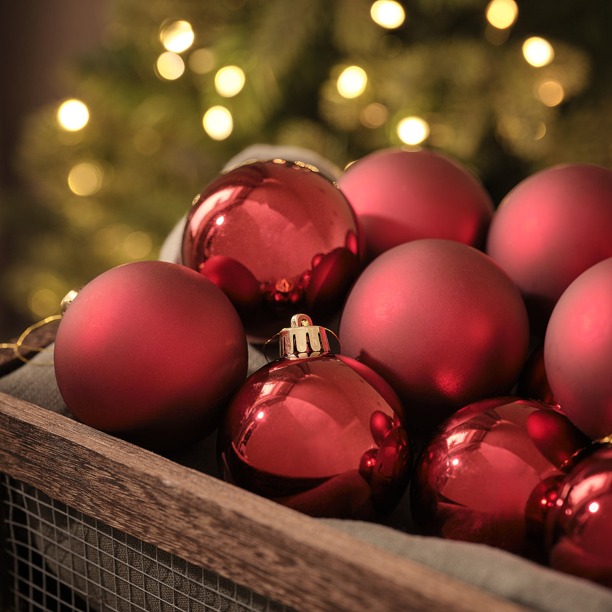 Rote Weihnachtskugeln in matter und glänzender Optik in Holzkiste vor Lichterglanz.