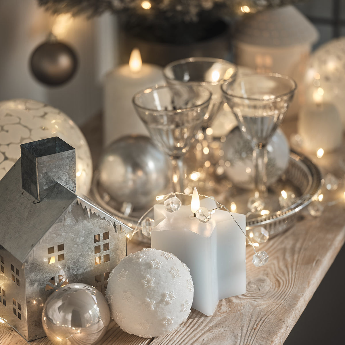Weiß-silberne Weihnachtsdekoration mit Kerzen und Häuschenfiguren auf Tisch.