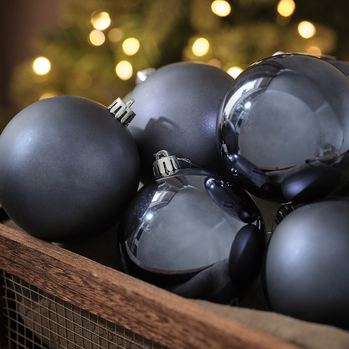 Blaugraue Weihnachtskugeln in matter und glänzender Optik in Holzkiste vor Lichterglanz.