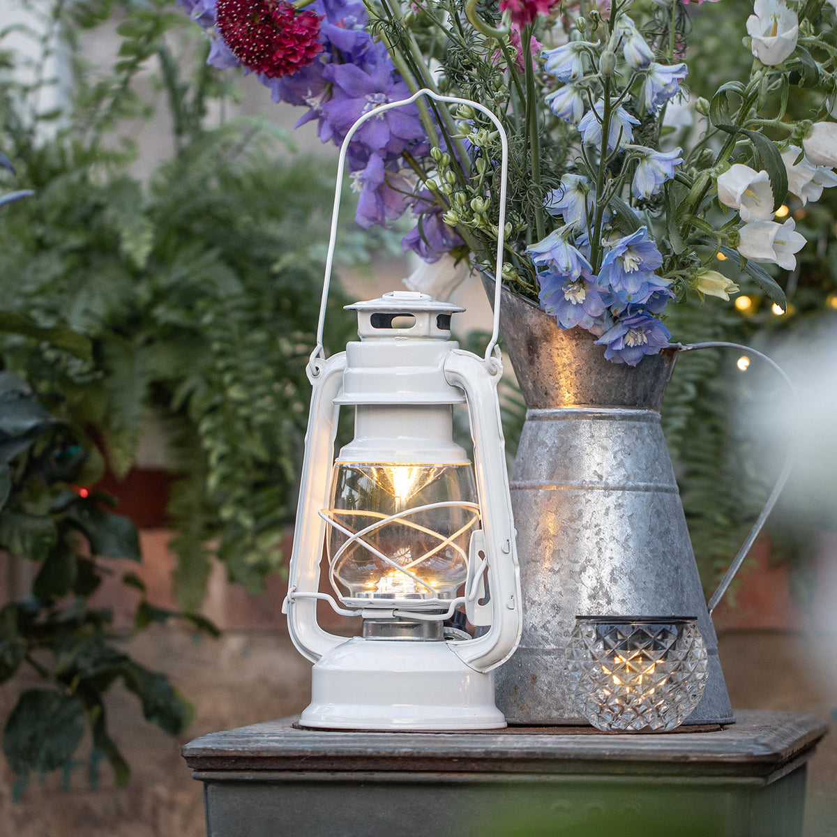 Weiße LED Sturmlaterne mit warmweißem Licht neben Metallkanne mit Blumen auf Holztisch