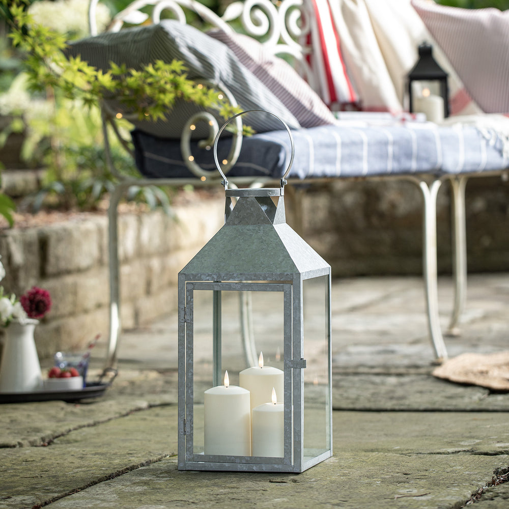 Große verzinkte Outdoor-Laterne mit drei TruGlow LED-Kerzen auf Steinboden vor Gartenbank