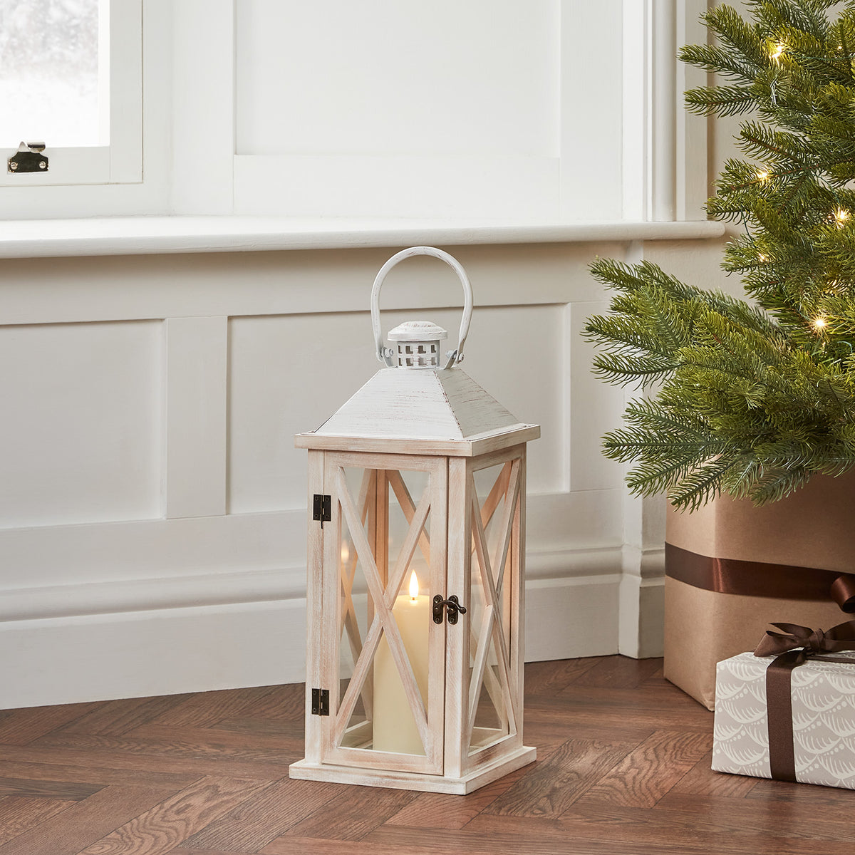 Weiße Holzlaterne mit TruGlow LED Kerze neben Weihnachtsbaum und Geschenken auf Holzboden