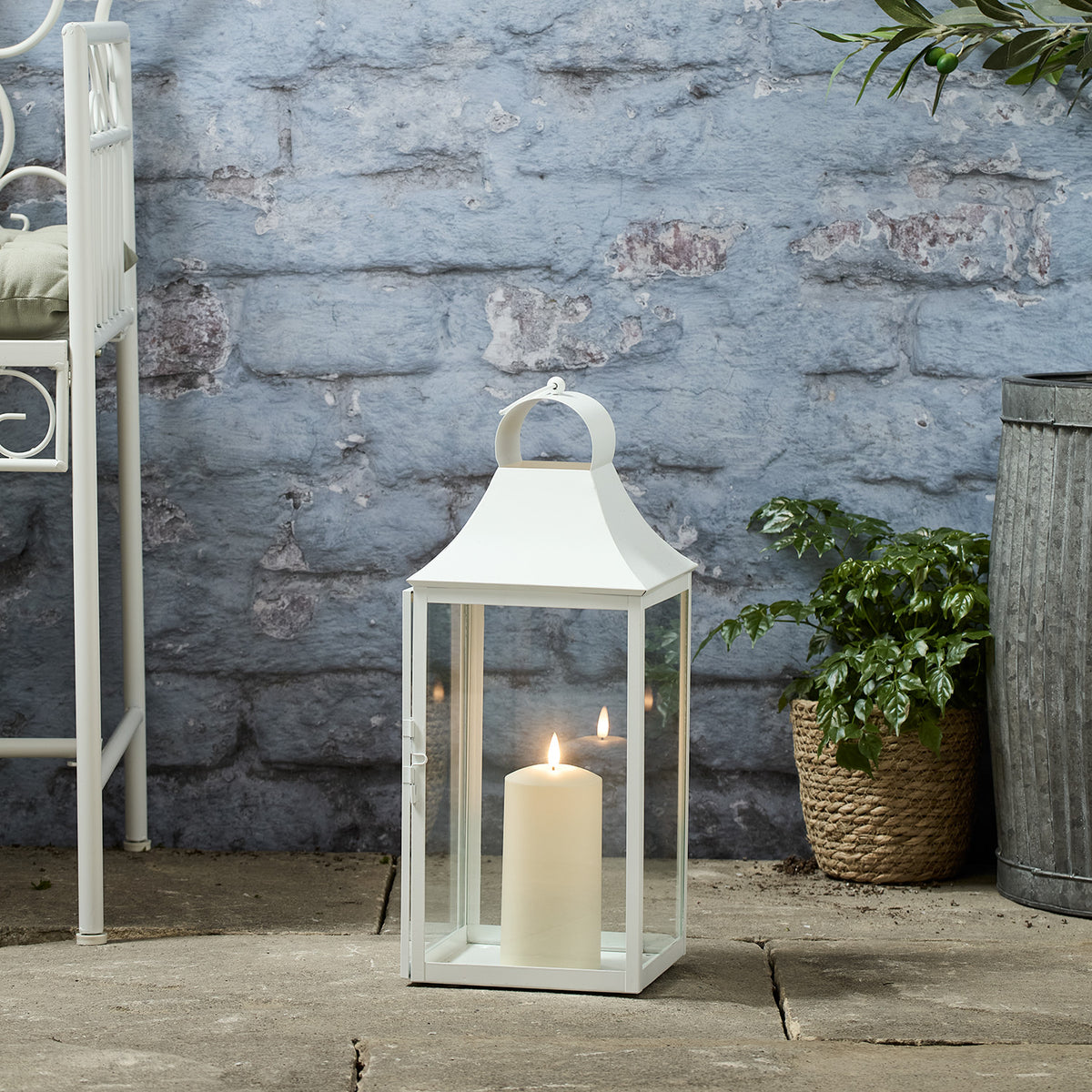 Weiße Gartenlaterne mit TruGlow® LED-Kerze auf Steinboden neben Pflanzen und Metalltopf