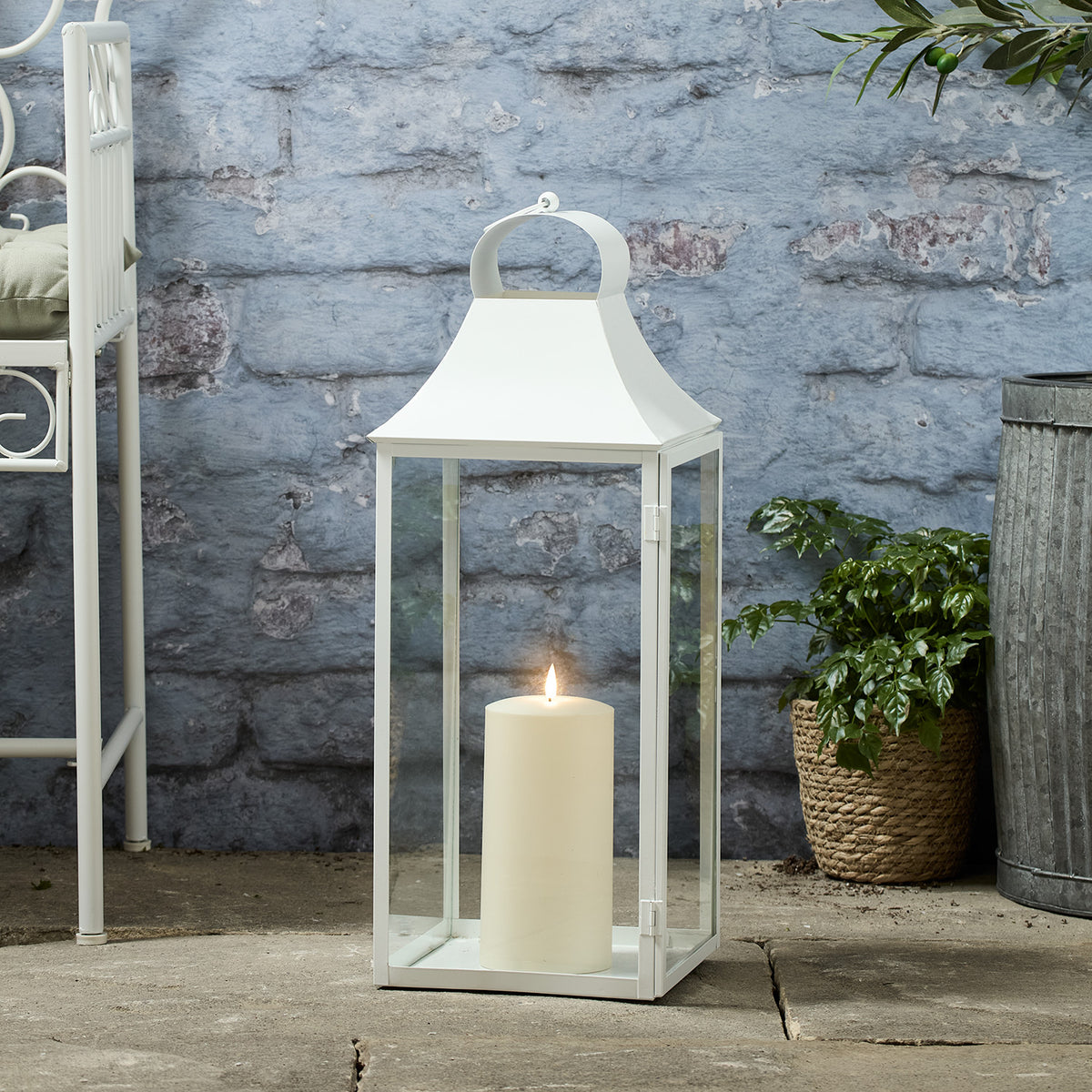 Große weiße Gartenlaterne mit leuchtender TruGlow® LED-Kerze vor blauer Wand und Pflanzen