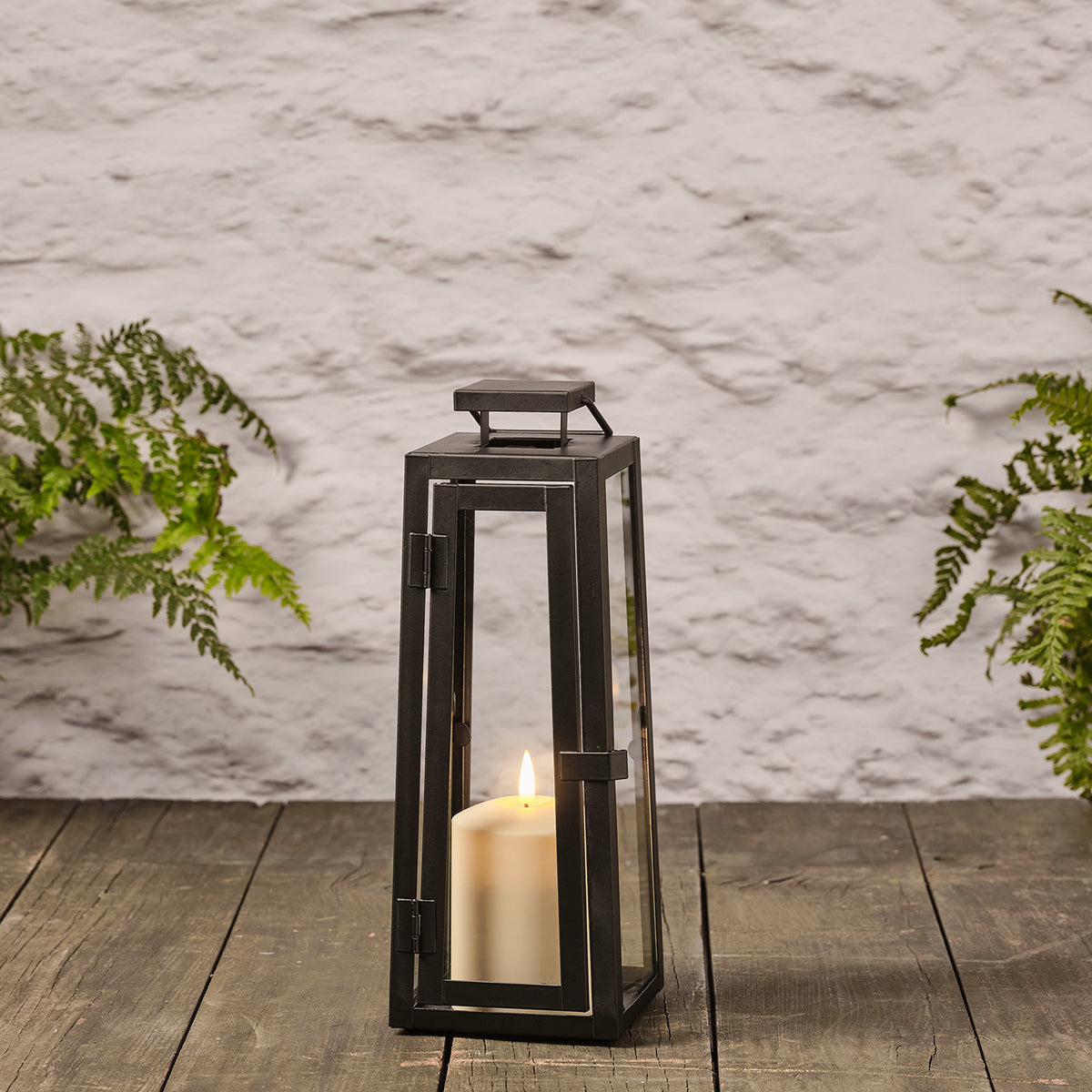 Schwarze Laterne mit Glas und TruGlow LED-Kerze auf Holzboden vor weißer Wand mit Farnen