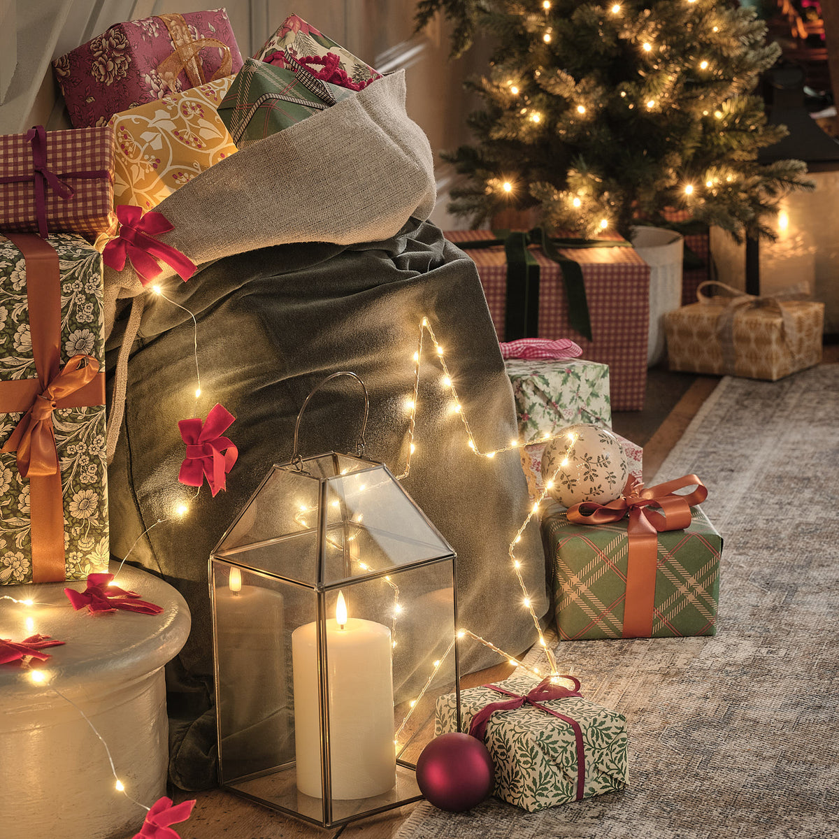 Goldene LED-Laterne mit flackernder Kerze und Lichterkette vor Weihnachtsgeschenken und Tanne
