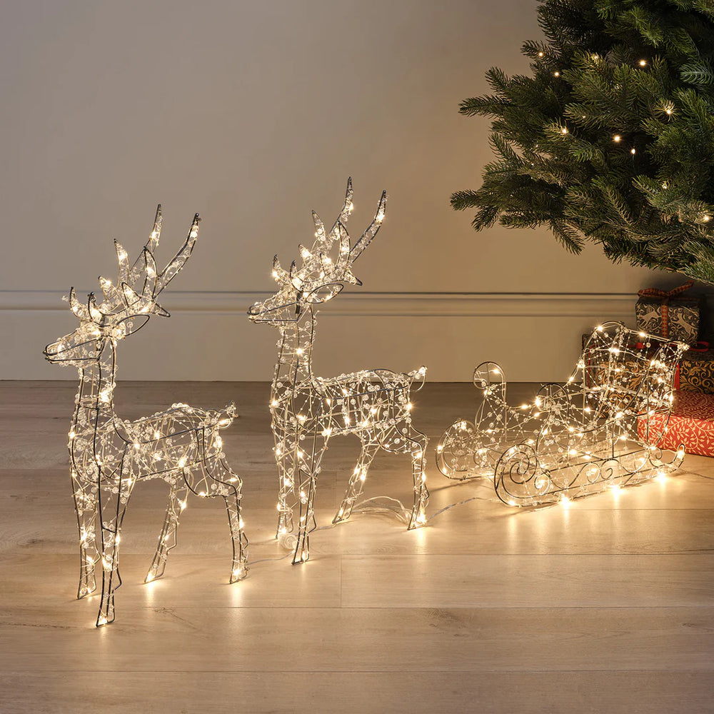 LED-Rentiere mit Schlitten aus Draht mit warmweißen Lichtern neben Weihnachtsbaum im Innenraum