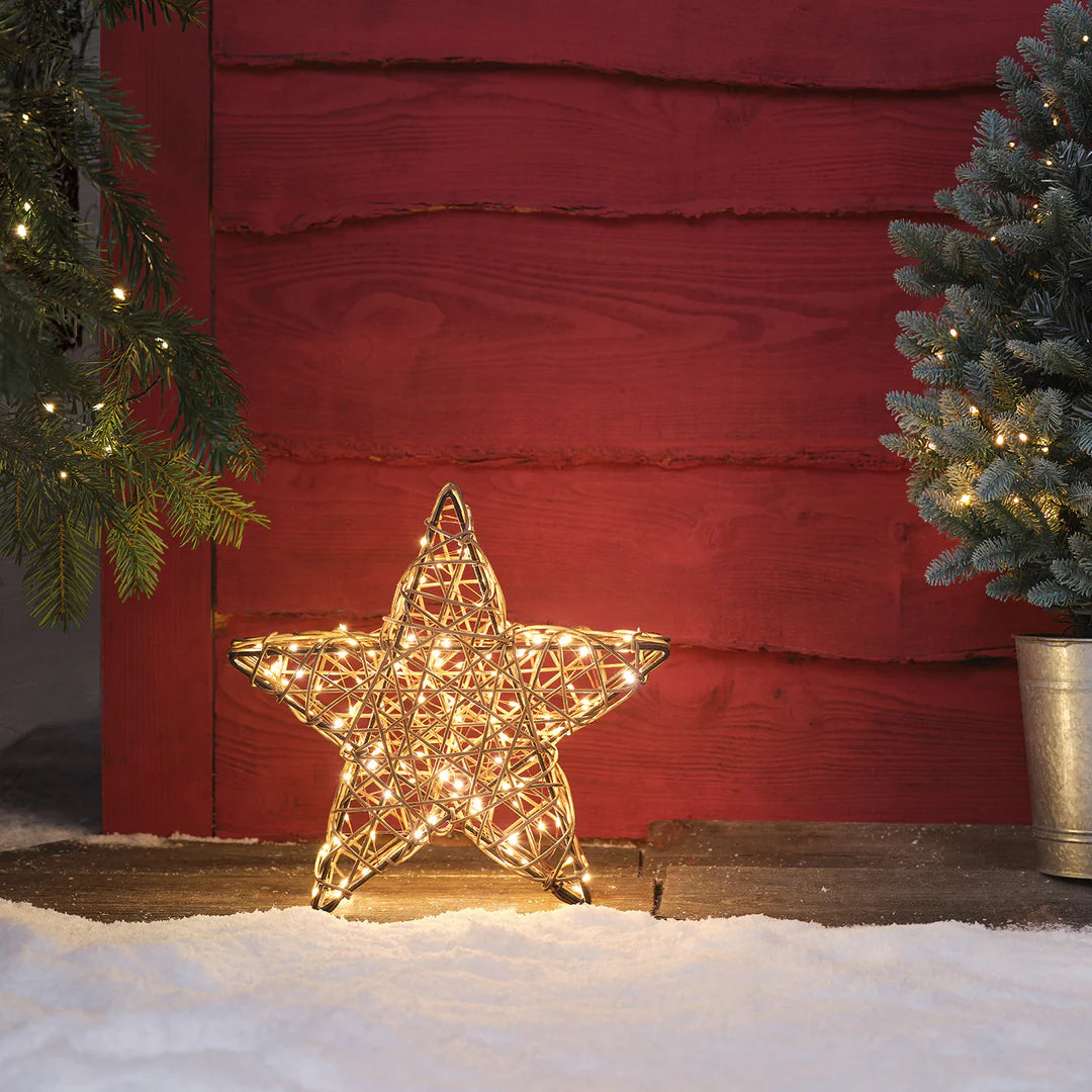 Rattan-LED-Stern mit warmweißen Lichtern auf Schnee vor roter Holzwand