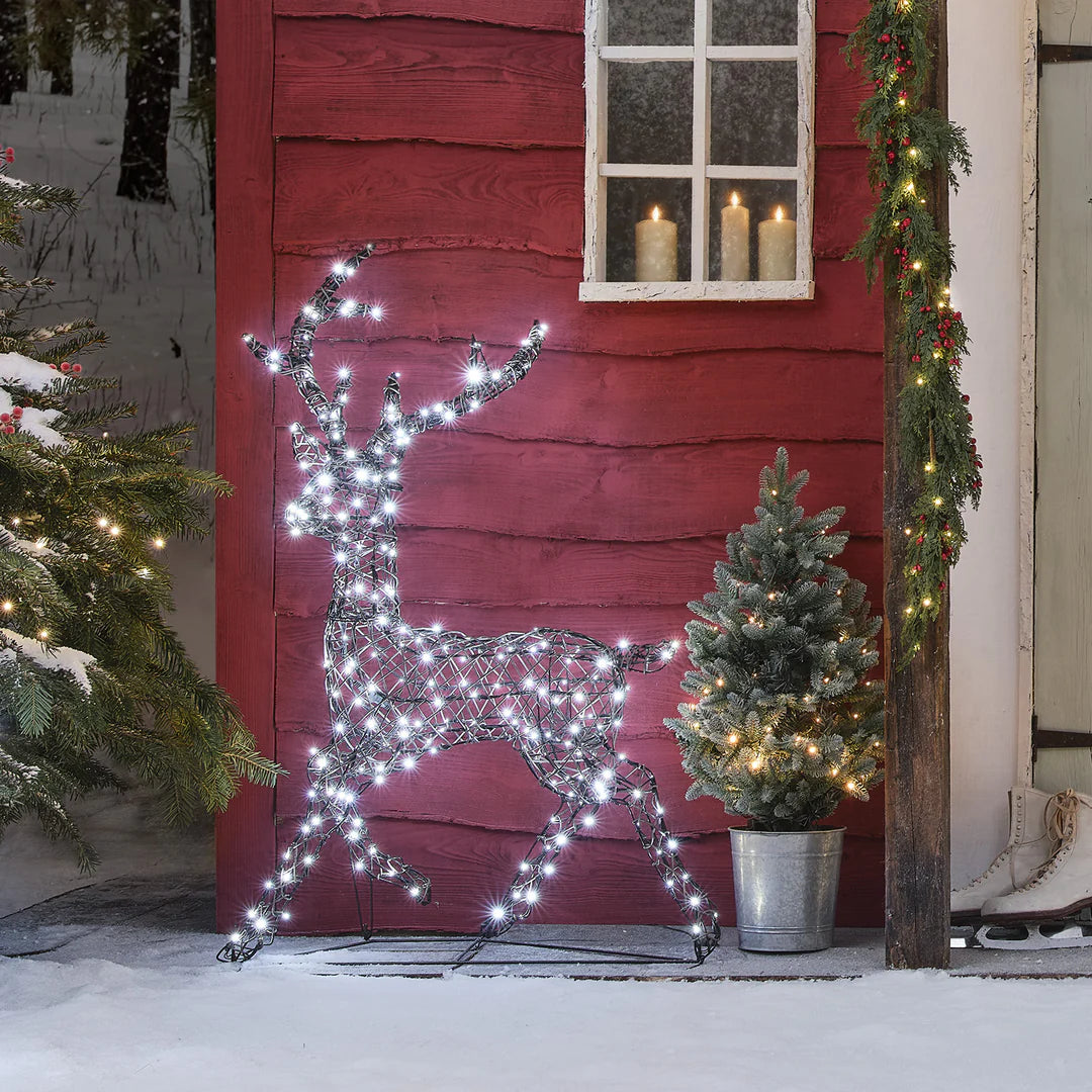 LED-Rentier mit kaltweißen Lichtern vor rotem Haus und Tannenbaum