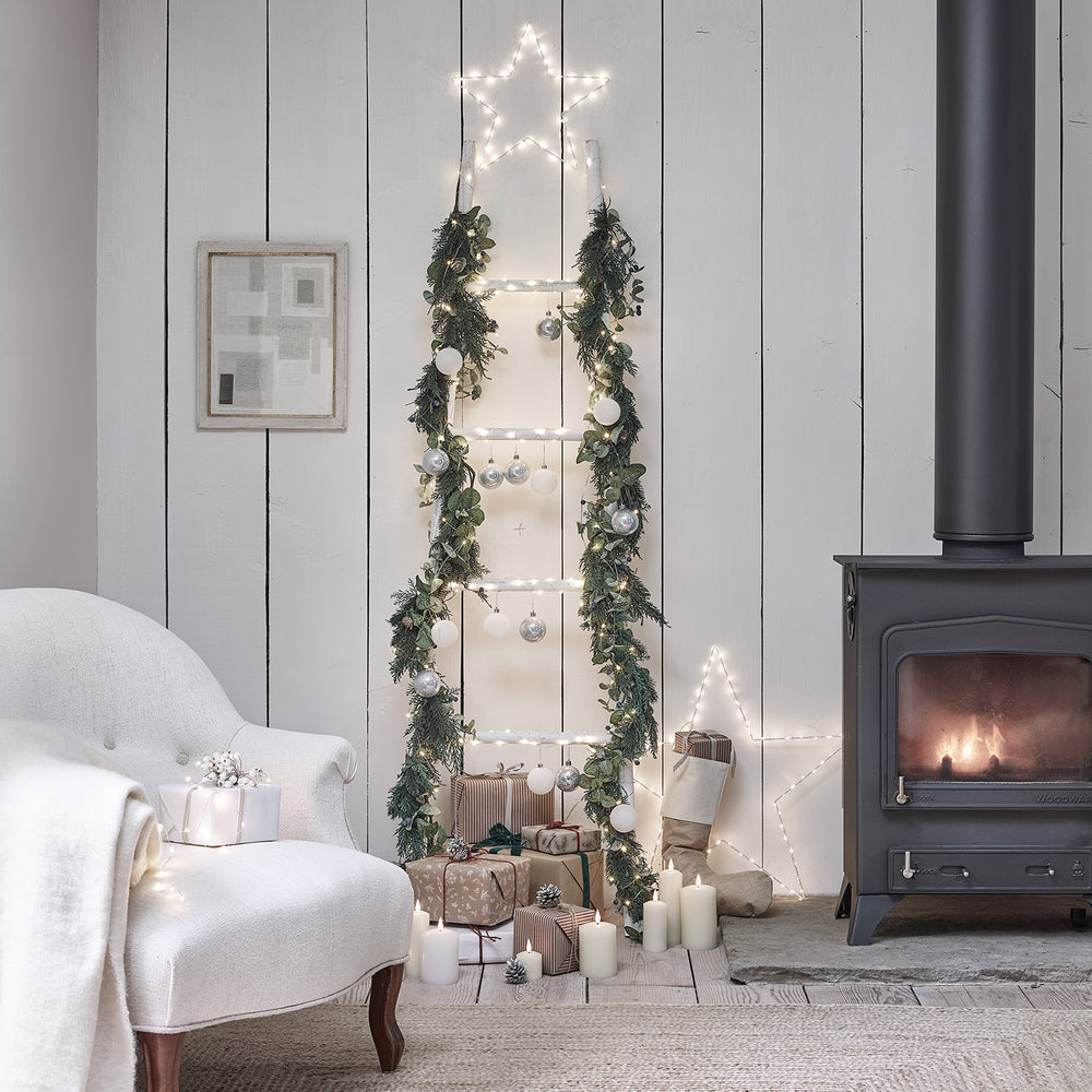 Weihnachtliche Dekoleiter mit Girlande, Kugeln und Stern-Lichterkette neben Kamin und Geschenken