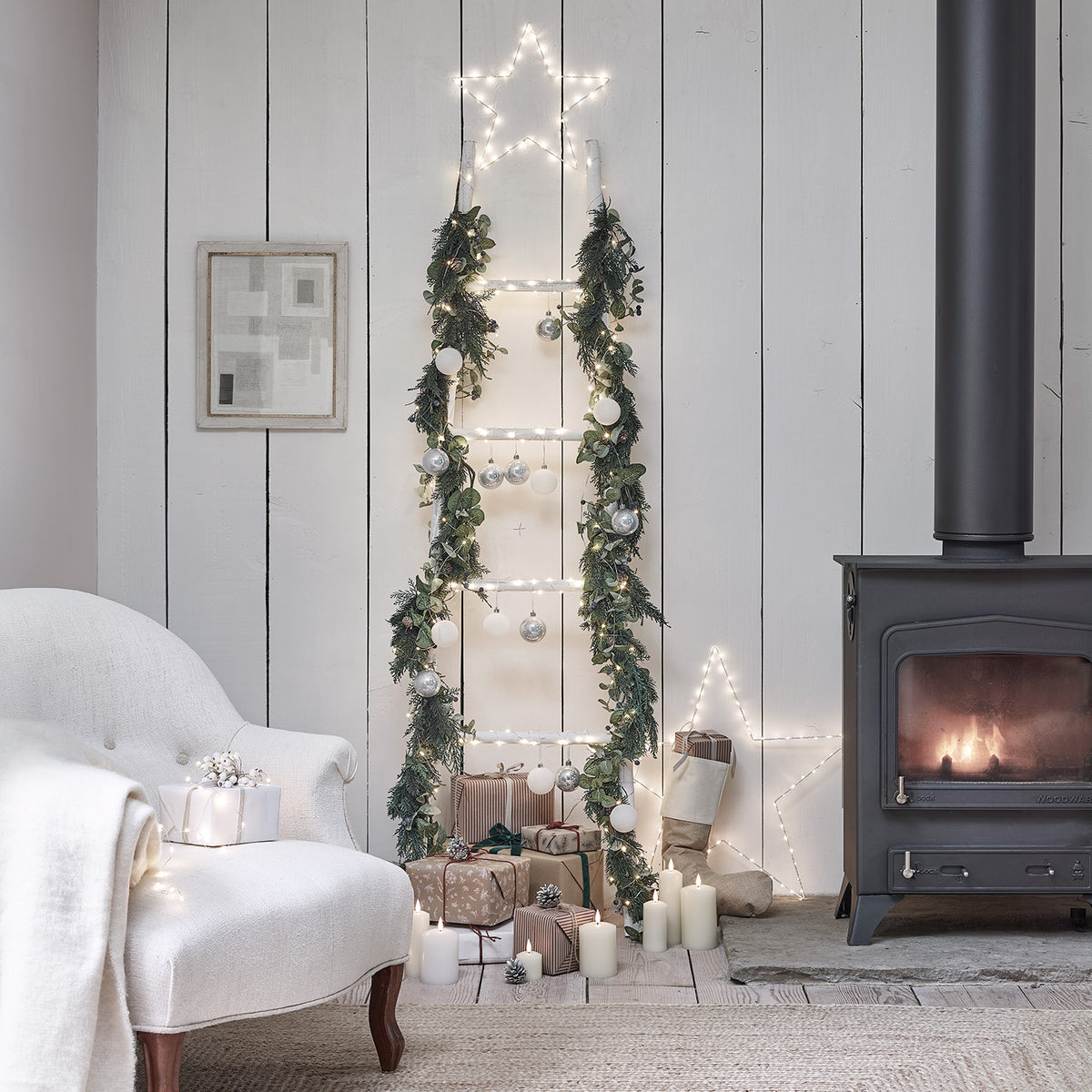 Weihnachtliche Dekoleiter mit Girlande, Kugeln und Stern-Lichterkette neben Kamin und Geschenken