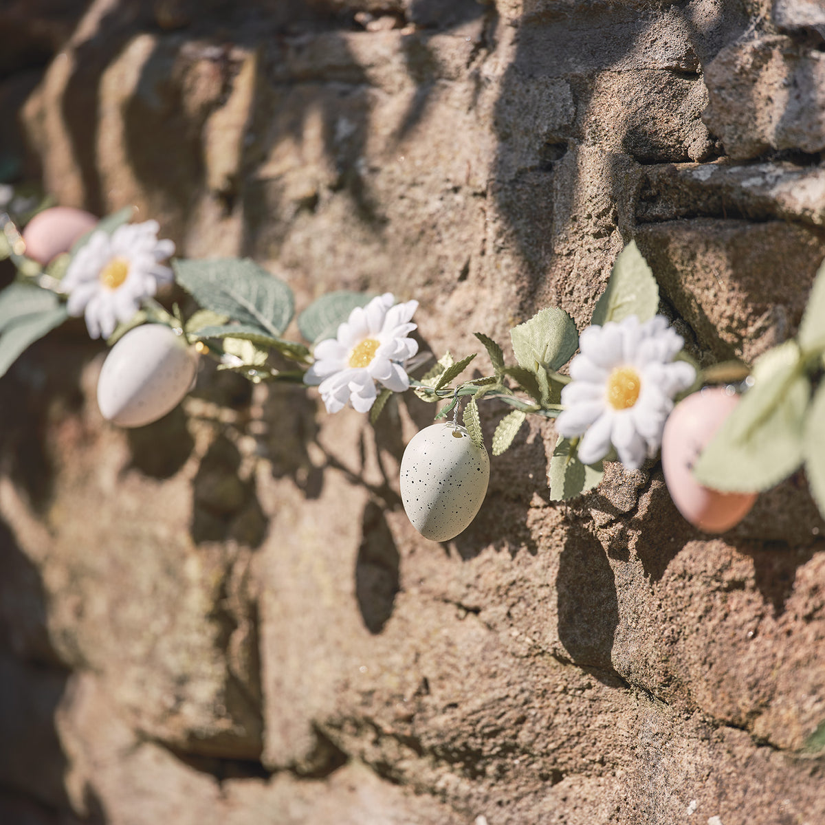 Girlande mit pastellfarbenen Ostereiern, Gänseblümchen und grünen Blättern an Steinmauer im Sonnenlicht