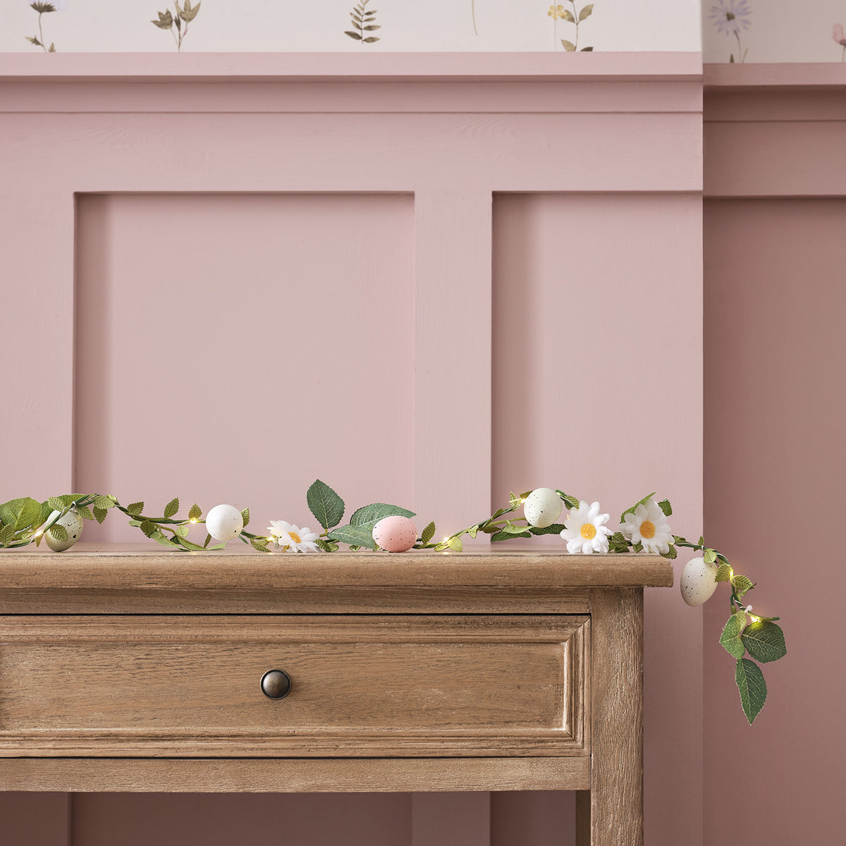Girlande mit Ostereiern, Gänseblümchen und Blättern auf Holzkommode vor rosa Wand