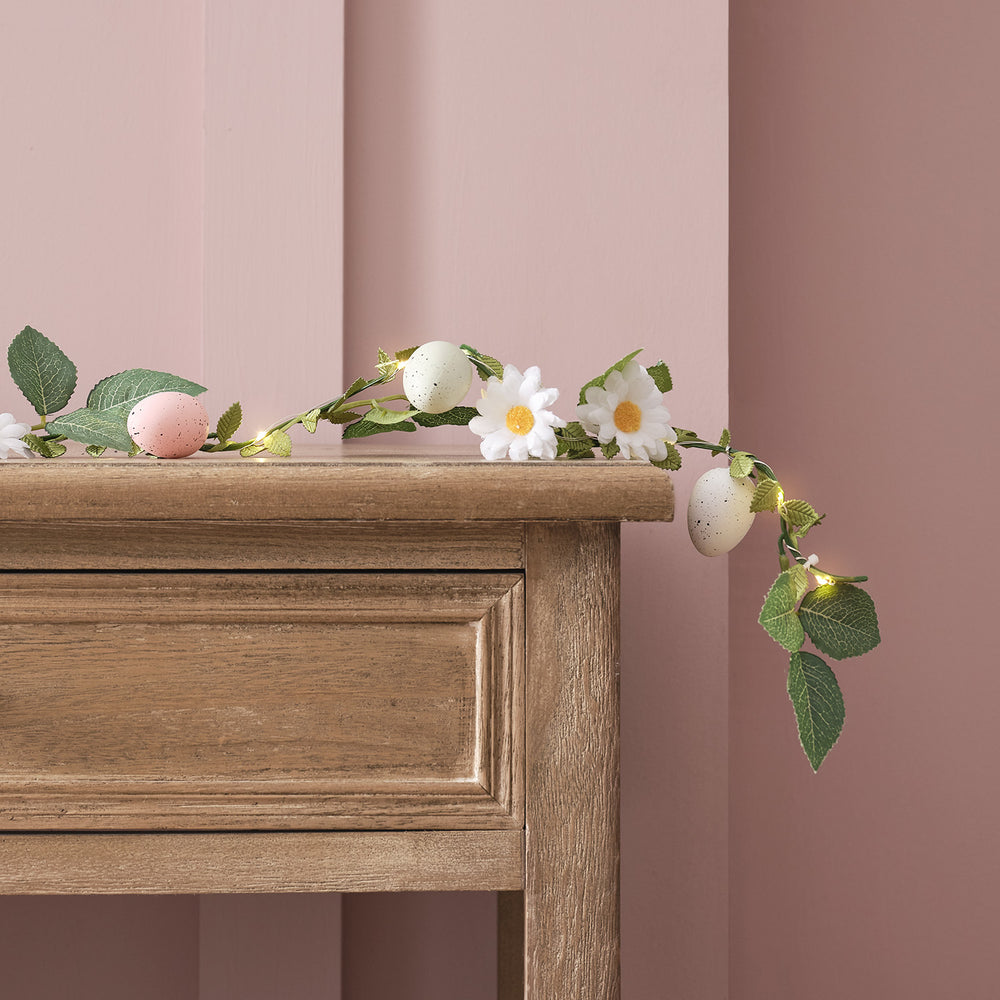 Oster-Girlande mit pastellfarbenen Eiern, Gänseblümchen und warmweißen LEDs auf Holzkommode