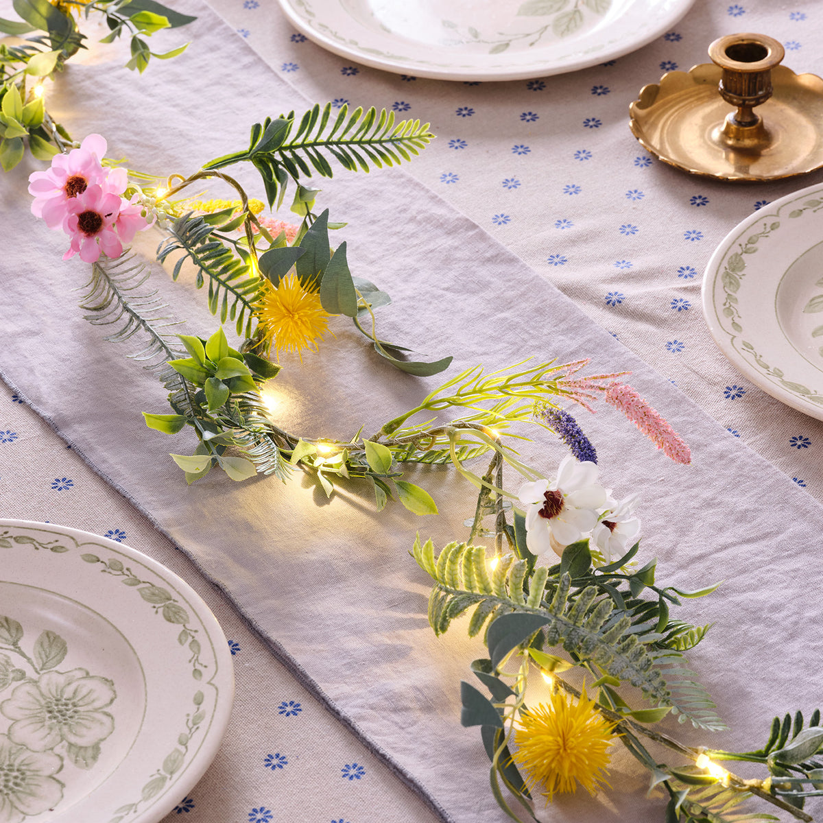 Frühlingsgirlande mit grünen Blättern, bunten Blumen und warmweißen LED-Lichtern auf einem gedeckten Tisch