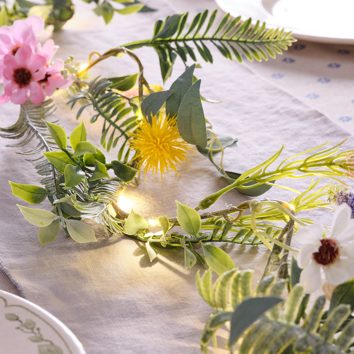 Frühlingsgirlande mit gelben und weißen Blumen sowie warmweißen LED-Lichtern auf einem hellen Stoff