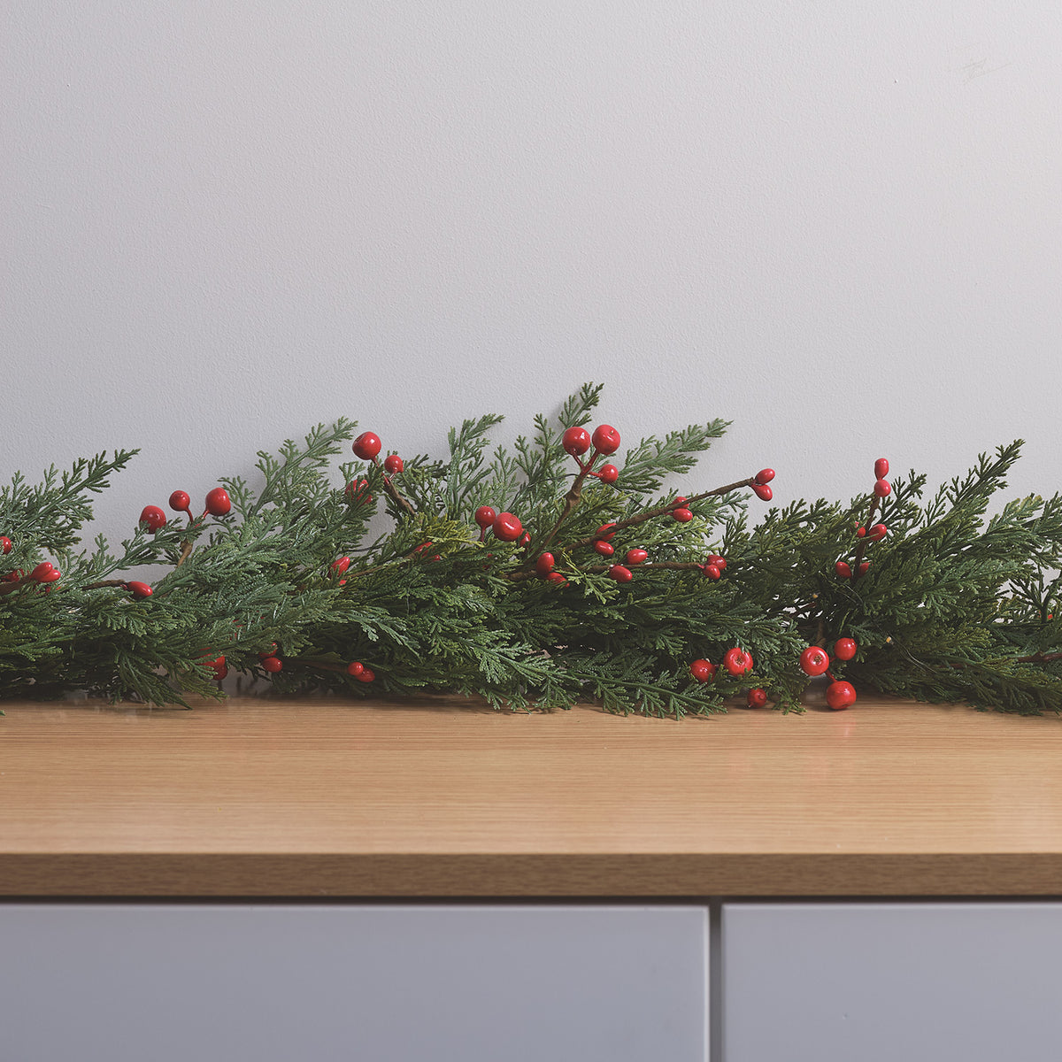 Lange Weihnachtsgirlande mit roten Beeren und LED-Lichtern auf Holztisch vor weißer Wand