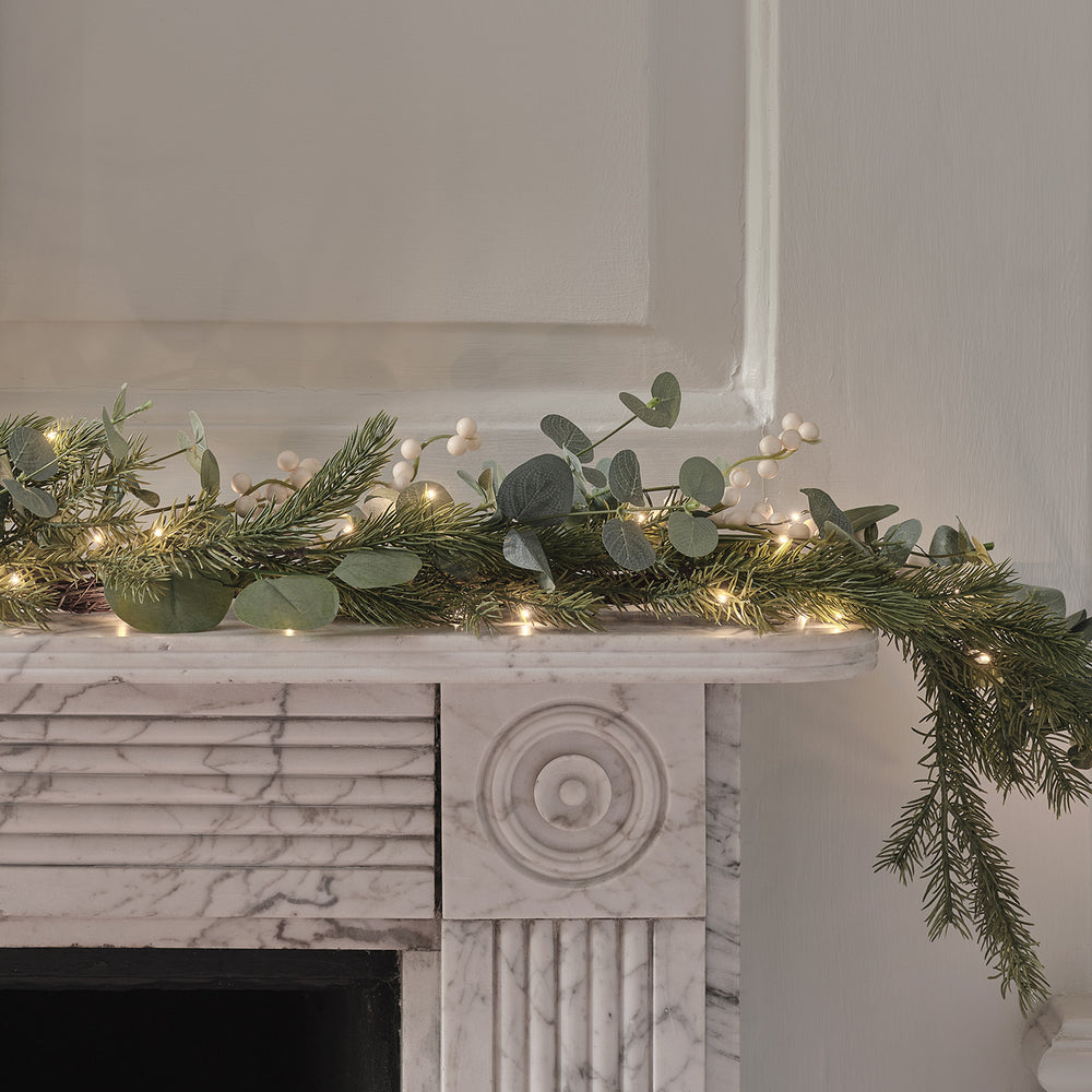 Beleuchtete Weihnachtsgirlande mit Eukalyptus und weißen Beeren auf weißem Kaminsims