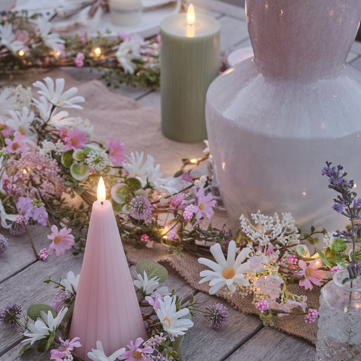 LED Blumengirlande mit pastellfarbenen Blüten neben einer rosa Kerze auf Holztisch