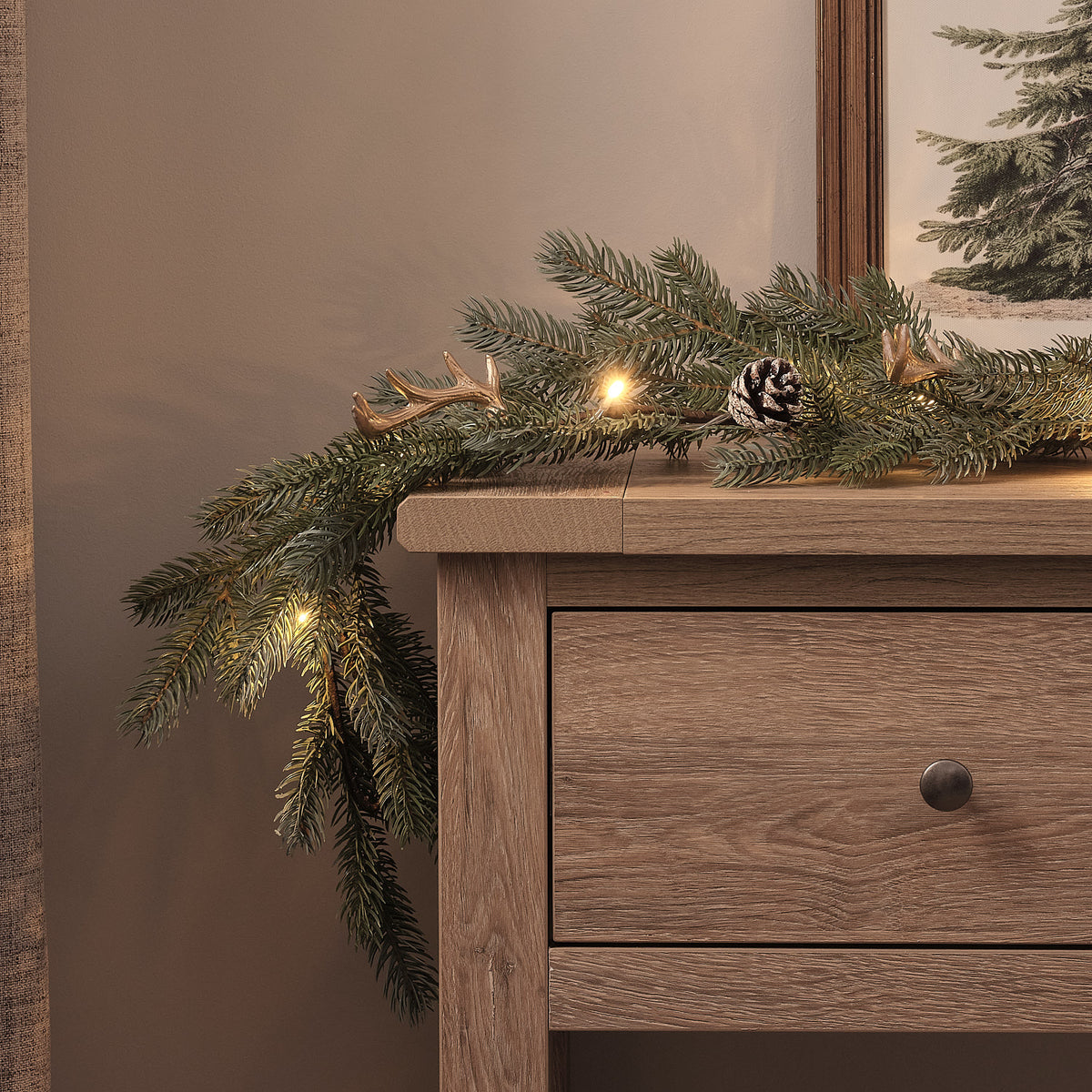 Detailaufnahme der LED-Weihnachtsgirlande mit künstlichem Laub, Geweih und Tannenzapfen auf Holzkommode