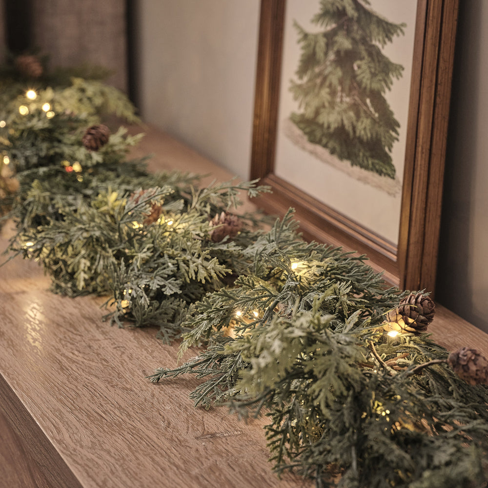 Beleuchtete Weihnachtsgirlande mit Tannenzapfen und warmweißen LEDs auf Holzkommode vor gerahmtem Bild