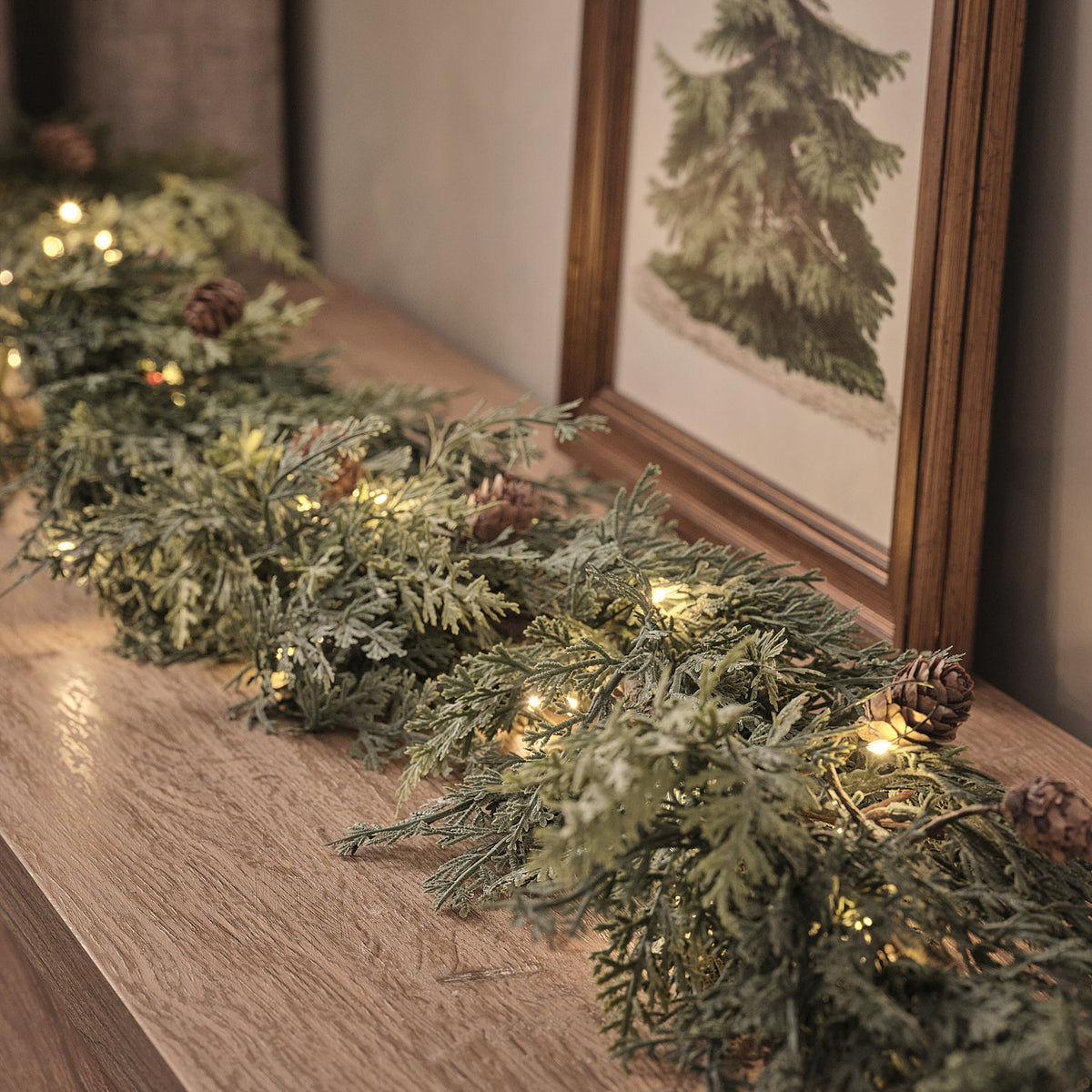 Beleuchtete Weihnachtsgirlande mit Tannenzapfen und warmweißen LEDs auf Holzkommode vor gerahmtem Bild