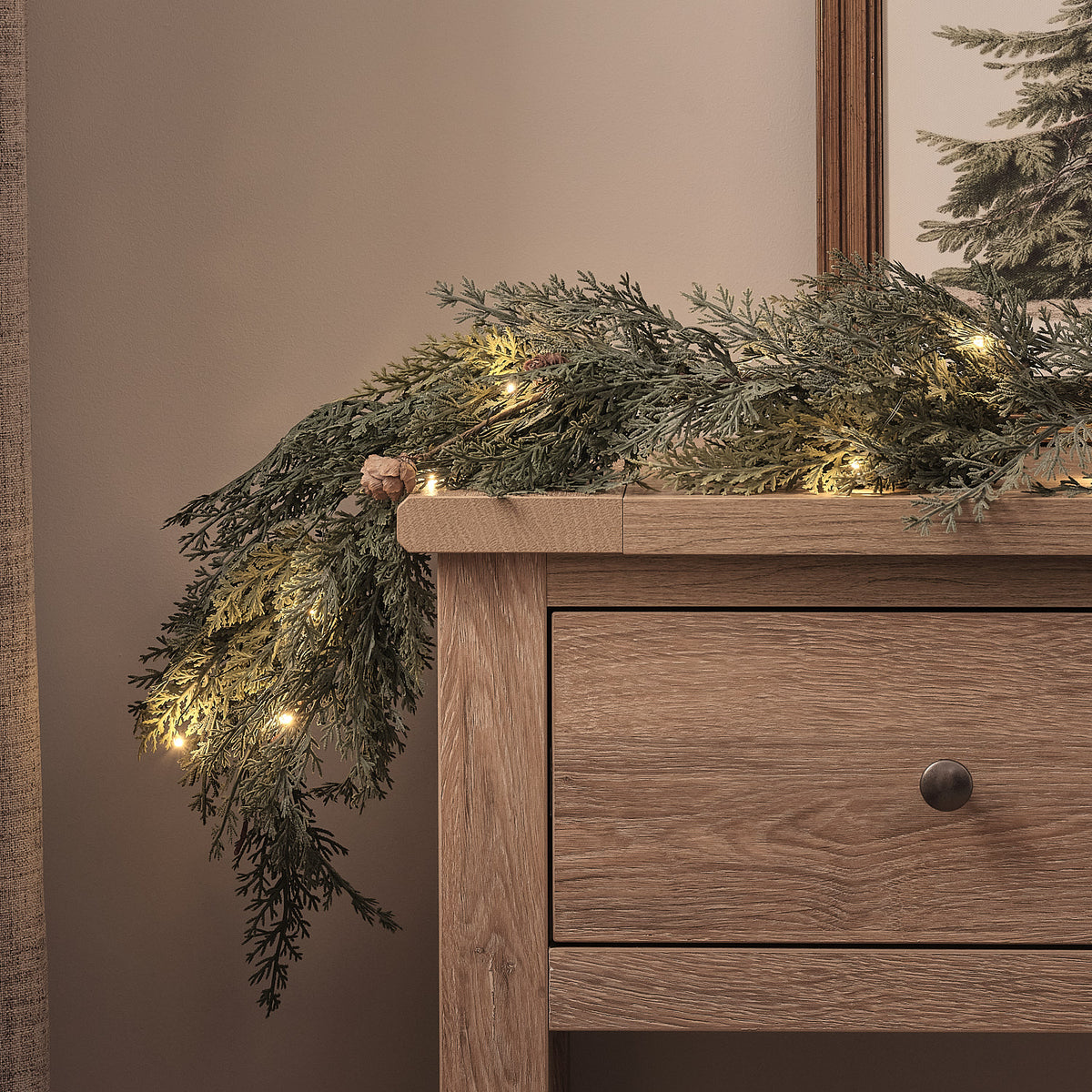 Beleuchtete Weihnachtsgirlande mit Tannenzapfen und warmweißen LEDs auf Holzkommode vor neutraler Wand