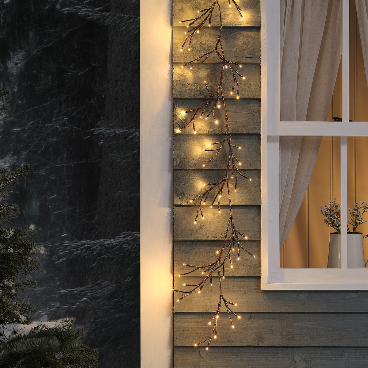 Beleuchtete LED-Zweiggirlande außen an Holzfassade neben Fenster mit Vorhang