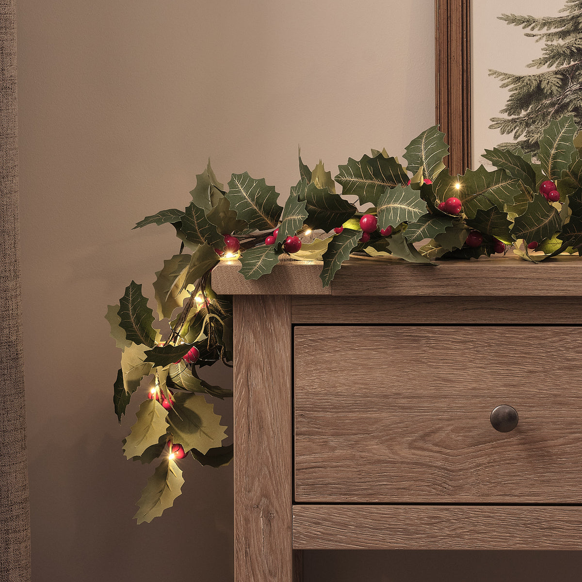 Beleuchtete Weihnachtsgirlande mit Stechpalmenblättern und roten Beeren auf Holzsideboard