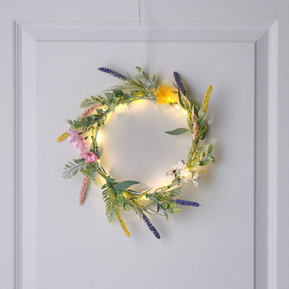 Beleuchteter Frühlingskranz mit Lavendel, rosa Anemonen und Löwenzahn an weißer Tür hängend