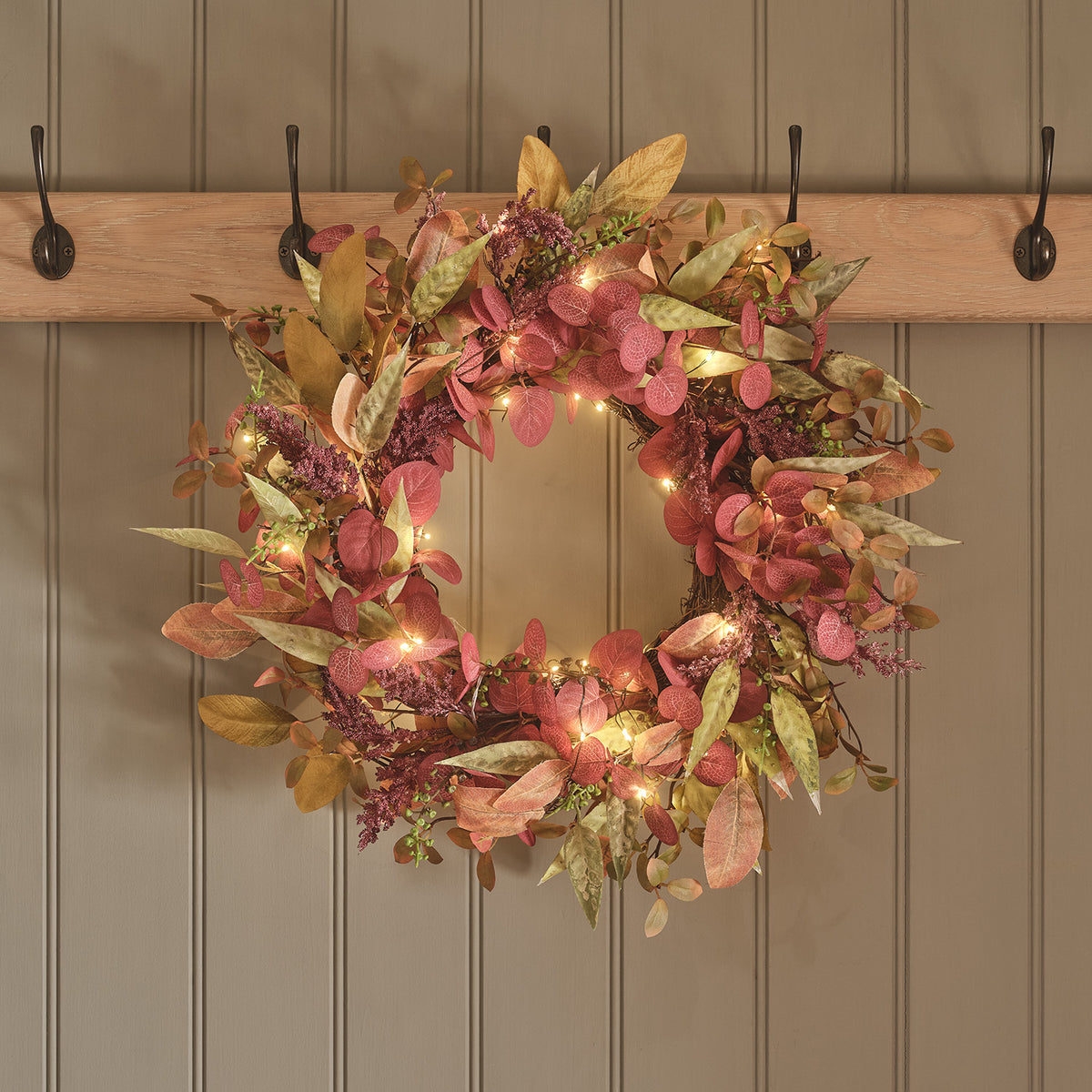 Herbstlicher Kranz mit künstlichen Blättern in Rot- und Grüntönen und warmweißen LEDs an einer Wand mit Haken