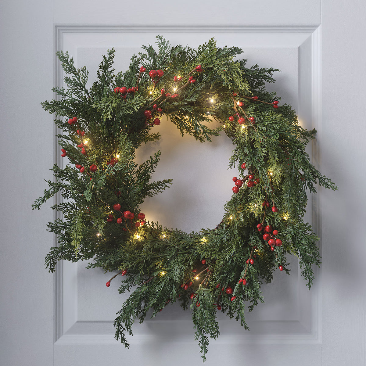 Weihnachtskranz mit roten Beeren und warmweißen LED-Lichtern an weißer Tür