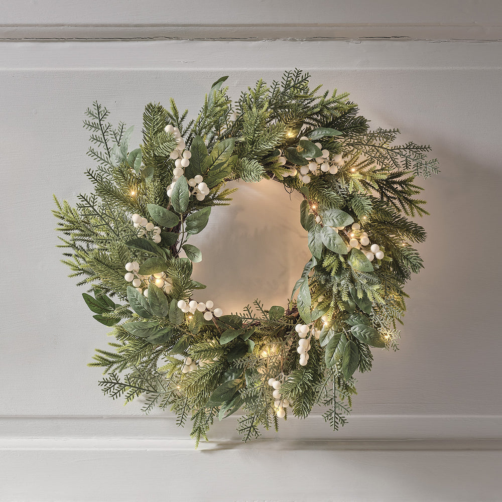 Weihnachtskranz mit grünen Blättern, weißen Beeren und warmweißen LED-Lichtern an weißer Wand