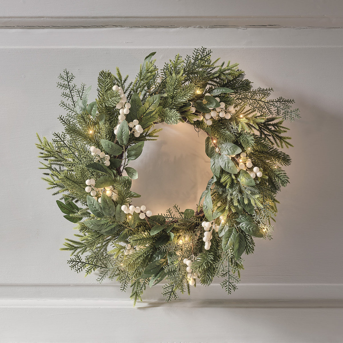 Weihnachtskranz mit grünen Blättern, weißen Beeren und warmweißen LED-Lichtern an weißer Wand