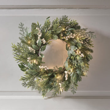 Weihnachtskranz mit grünen Blättern, weißen Beeren und warmweißen LED-Lichtern an weißer Wand