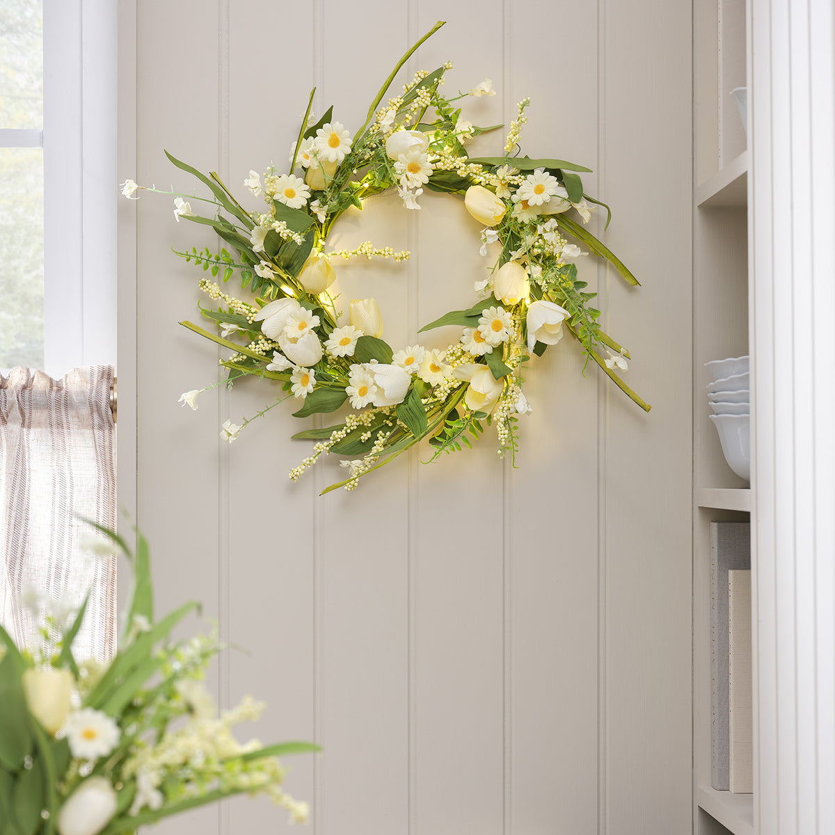 Frühlingskranz mit gelben Tulpen und weißen Gänseblümchen an heller Wand neben Fenster und Regal