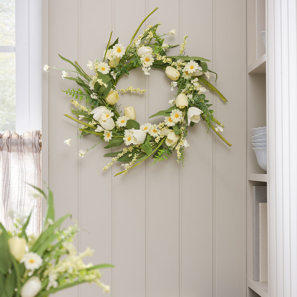 Frühlingskranz mit gelben Tulpen und weißen Gänseblümchen an heller Wand neben Fenster und Regal