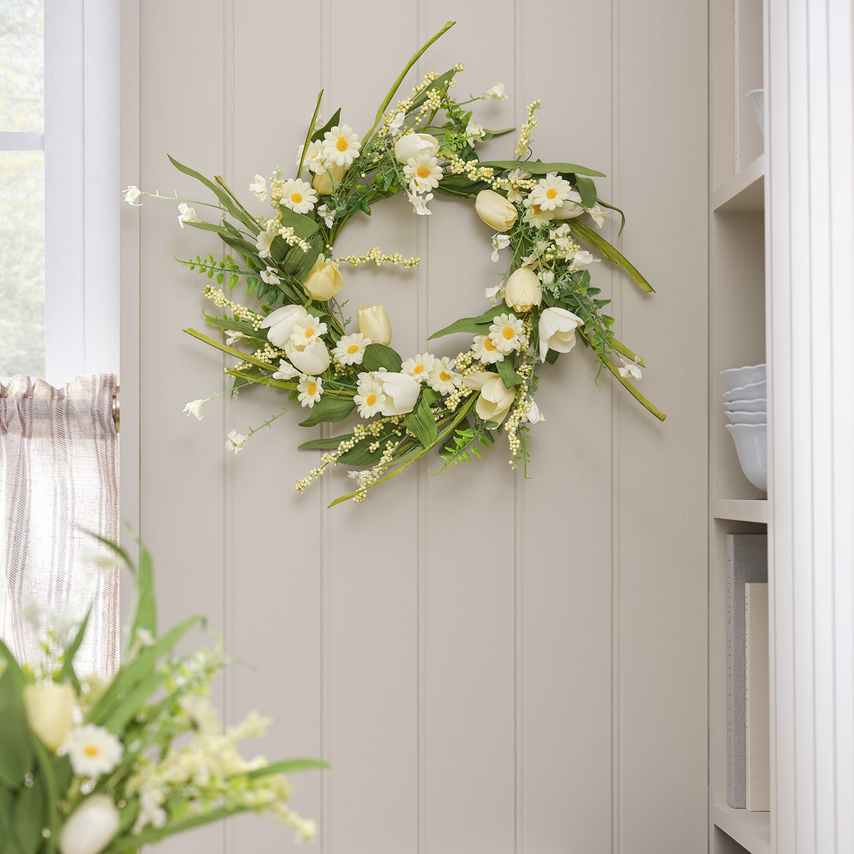 Frühlingskranz mit gelben Tulpen und weißen Gänseblümchen an heller Wand neben Fenster und Regal