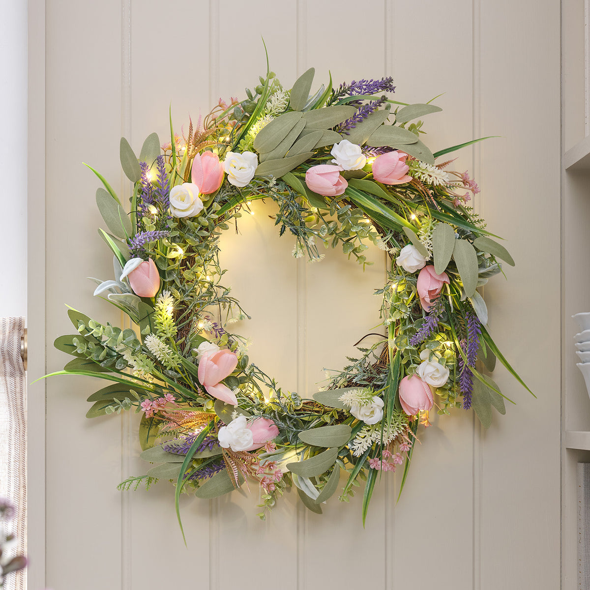 LED-Tulpenkranz mit rosa Tulpen, Lavendel, weißen Rosen und warmweißen Mikro-LEDs an einer hellen Tür